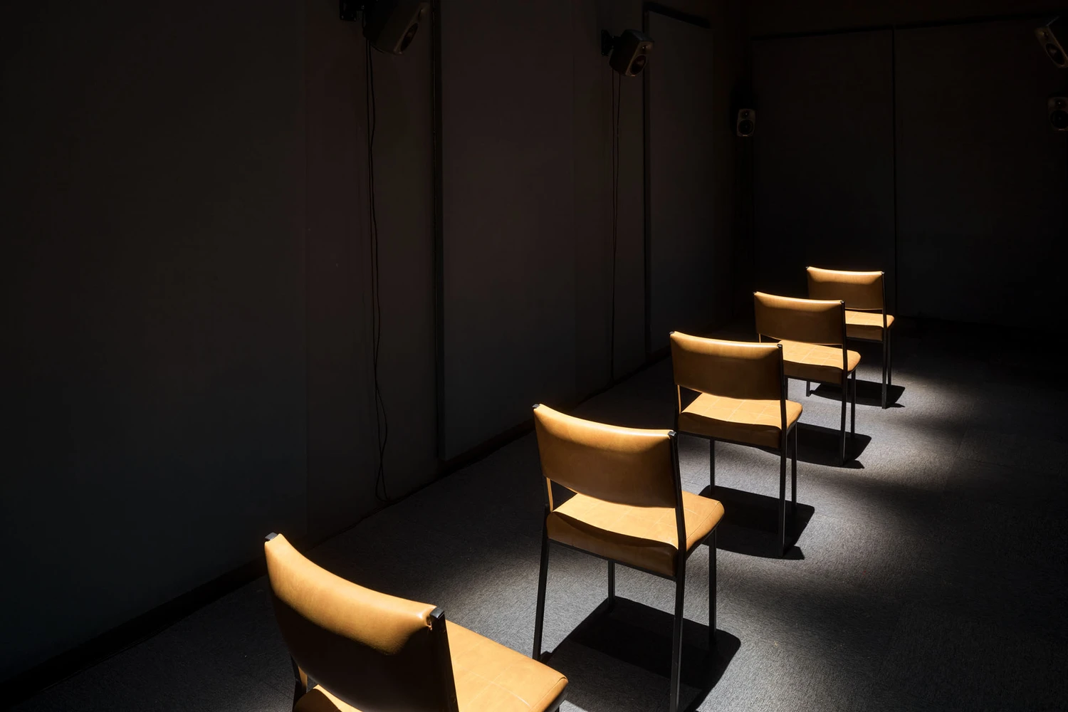 Five empty chairs are arranged in a line, facing forward. The walls of the room are blank, shadowed and unadorned, except for three speakers mounted close to the ceiling. Black cords run down from the speakers to the floor, camouflaged by darkness. The floor is paved with nondescript carpet tiles, like an office or waiting room.